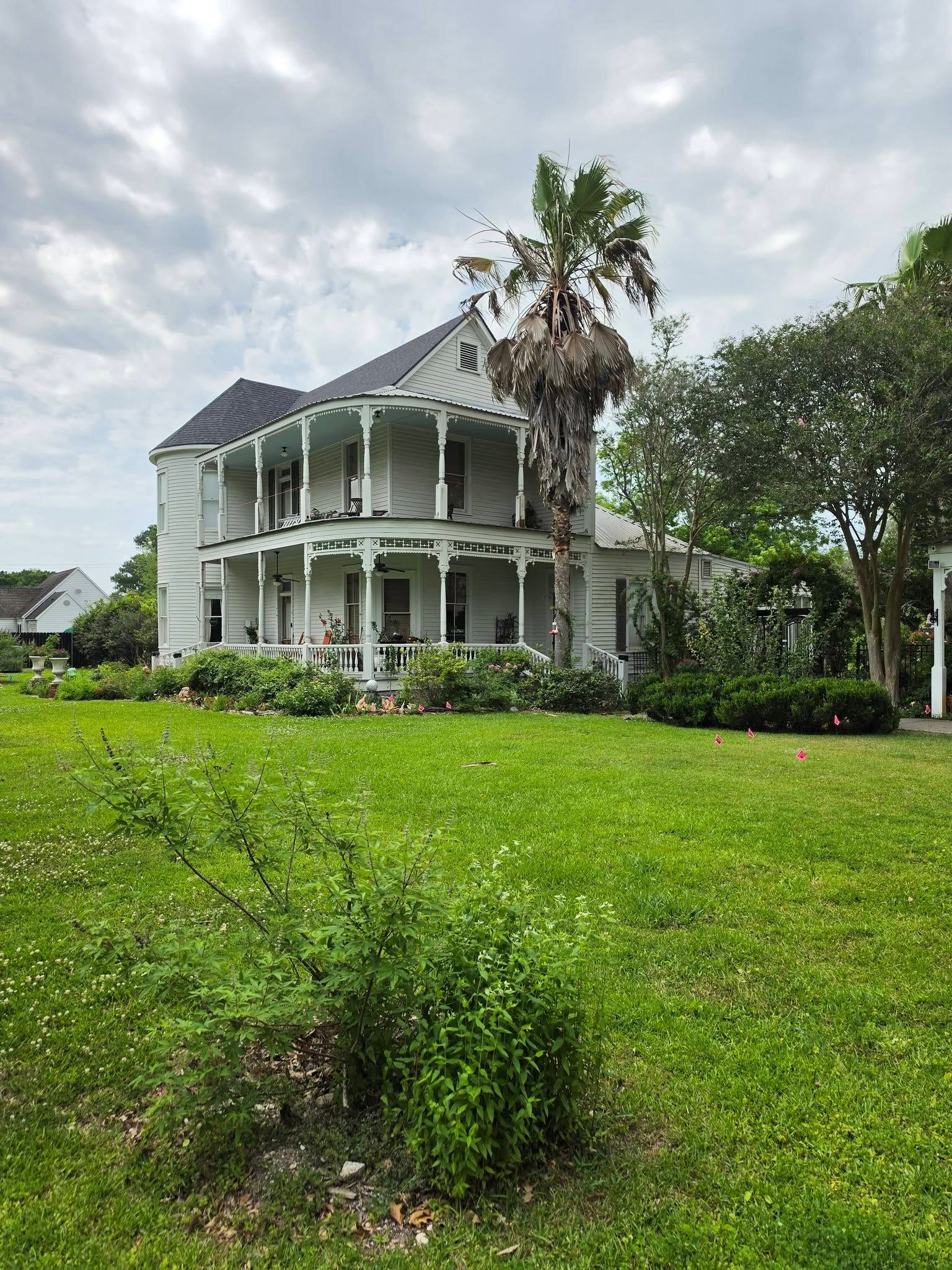 White two-story house with a wraparound porch beside a palm tree and green lawn under cloudy skies