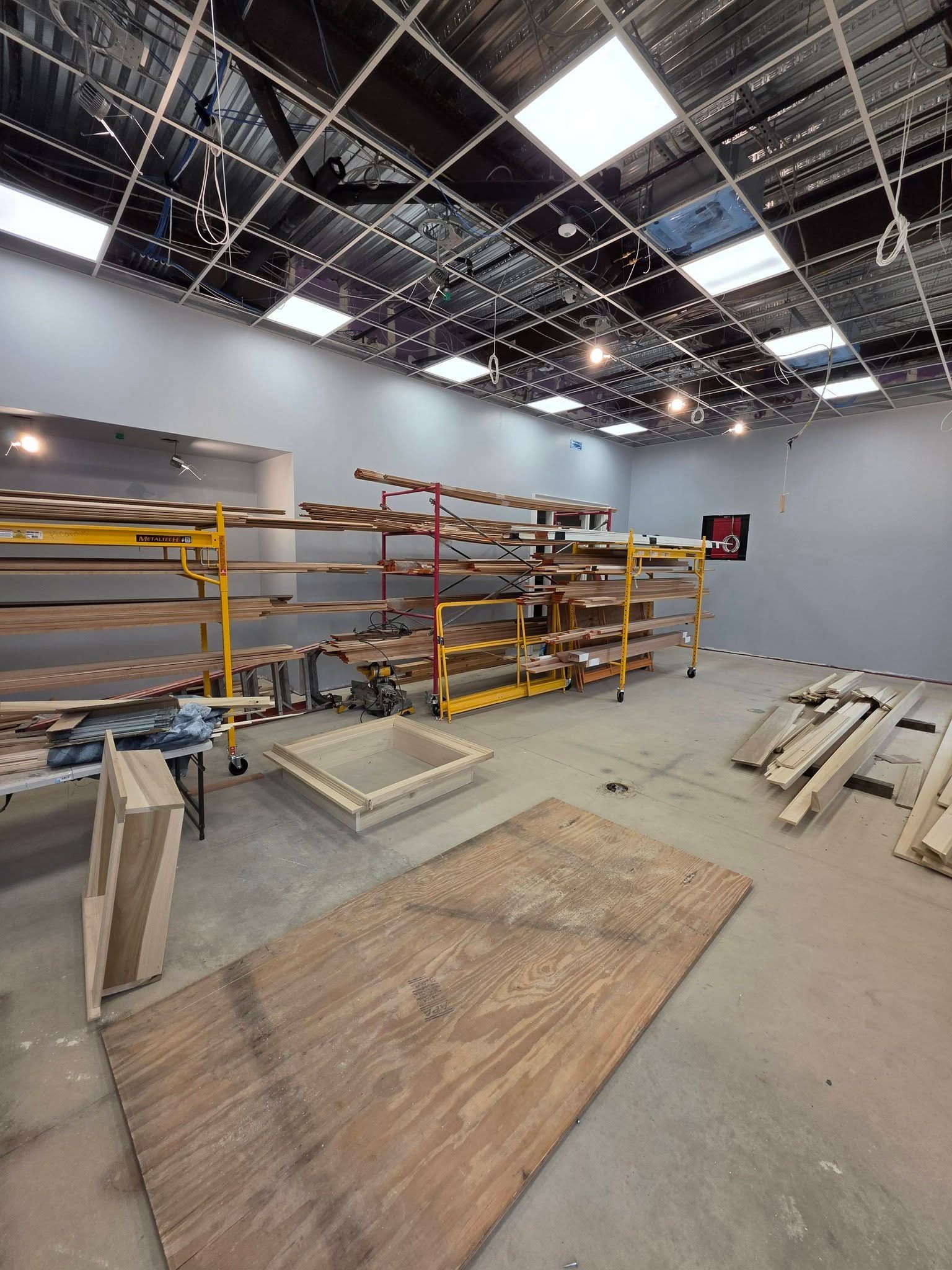Empty warehouse interior with scaffolding, shelves, wood panels, and a grid-lit ceiling under construction