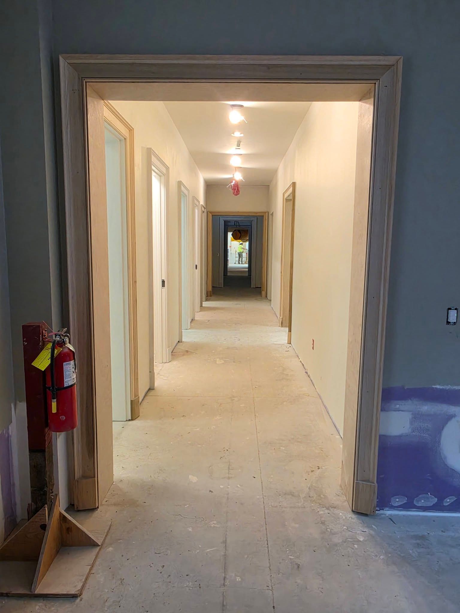 Empty hallway under renovation with open doorways, exposed walls, and a red fire extinguisher on the left.