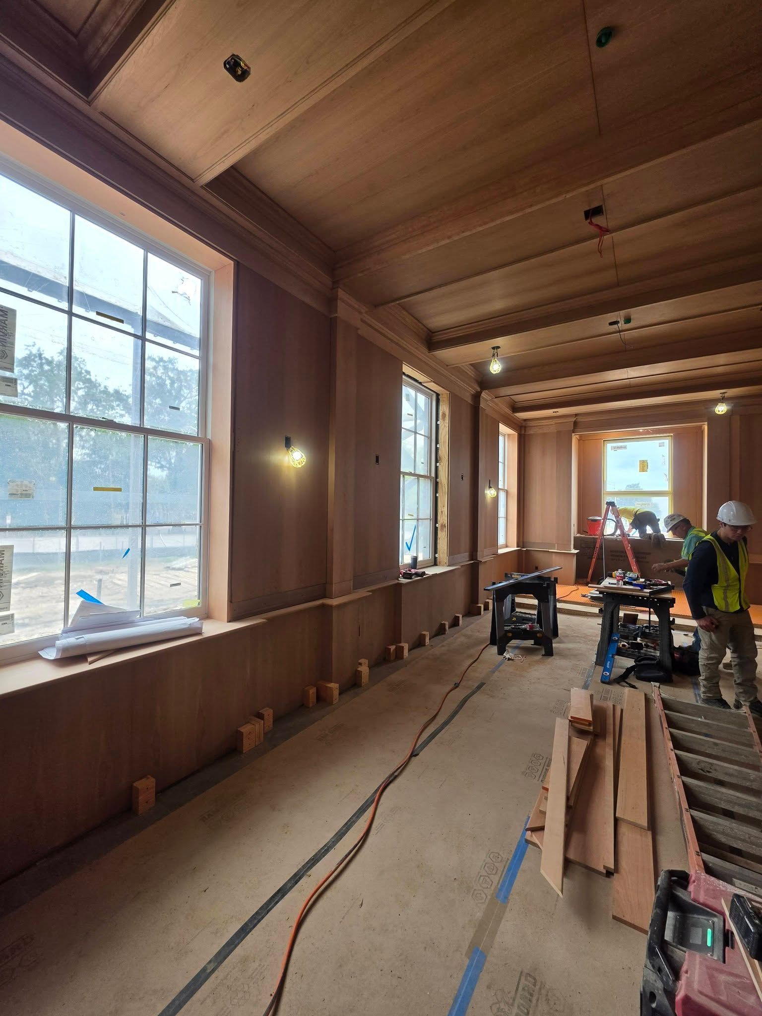 Wood-paneled interior under construction with workers, tools, and large windows letting in daylight.