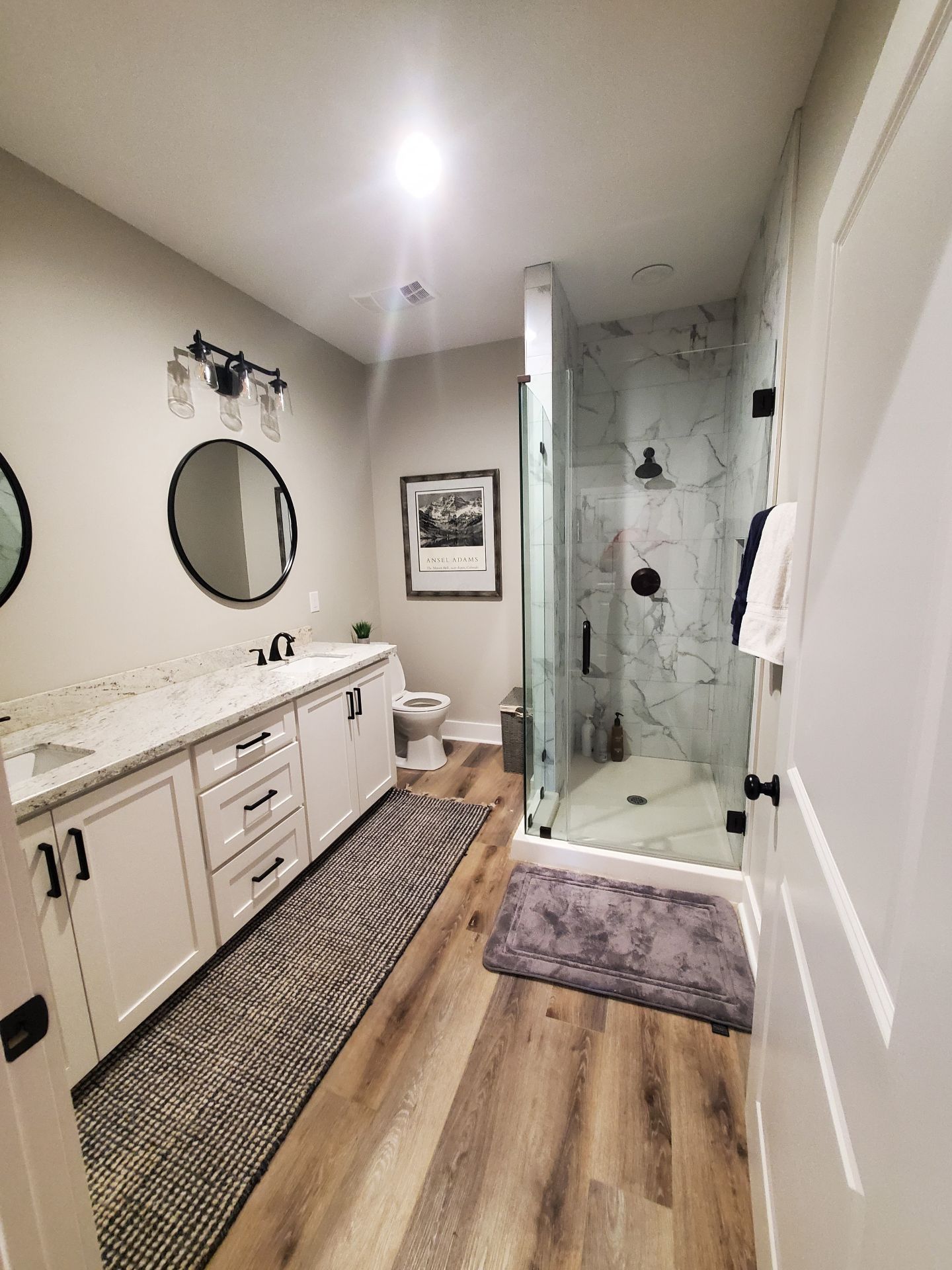 Modern bathroom with double vanity, round mirrors, glass shower, and wood-look floor tiles.