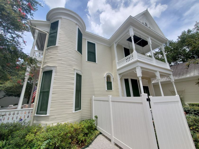 White Victorian-style house with a rounded tower, green shutters, and a screened porch behind a white fence