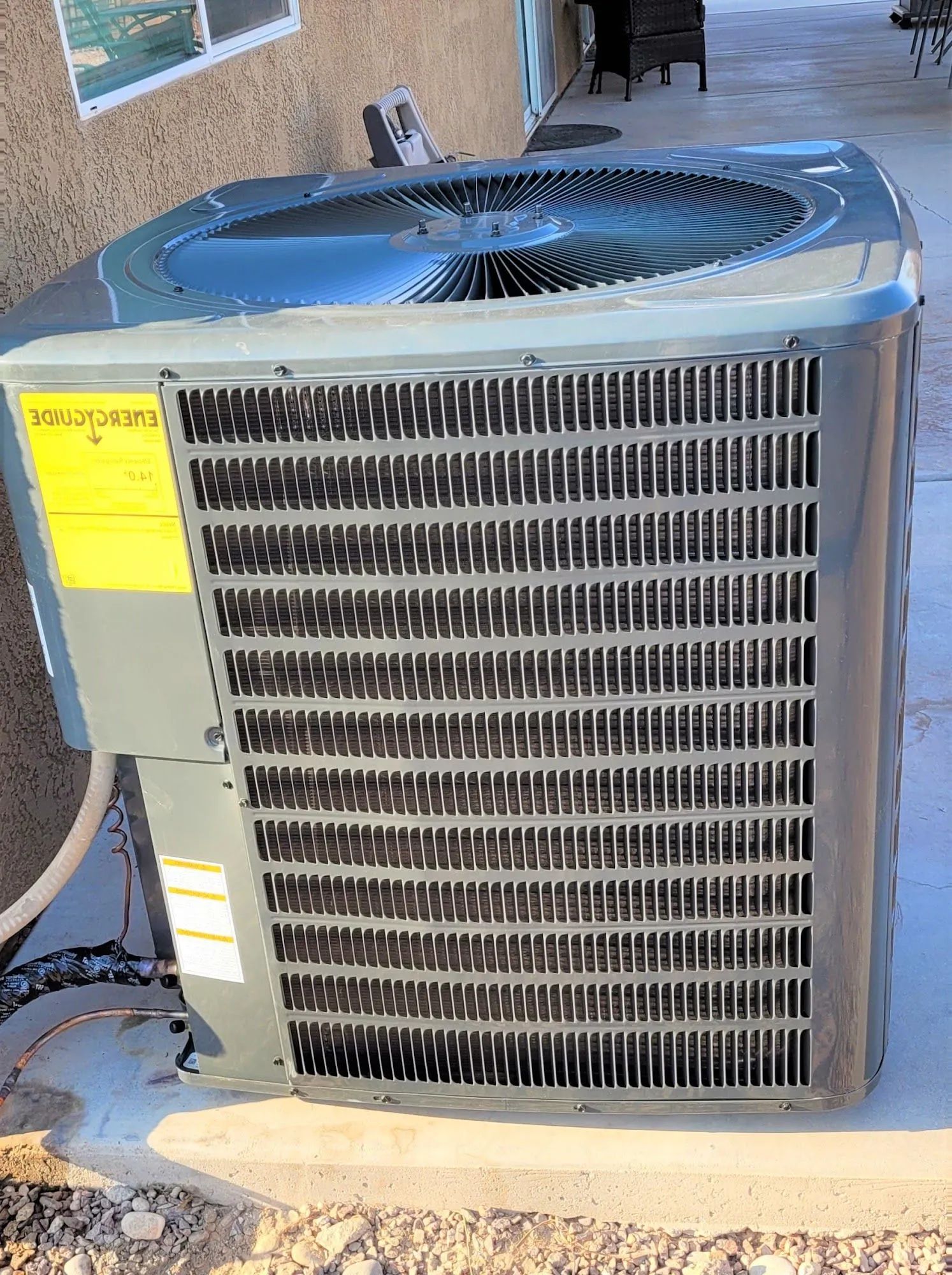Outdoor air-conditioning unit next to a brick wall, with a fan on top and yellow label on the side