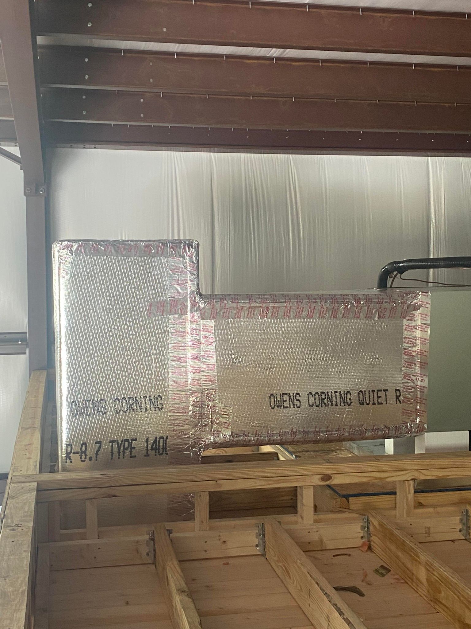 A foil-wrapped rectangular HVAC duct system mounted above wooden floor joists in an unfinished indoor space.