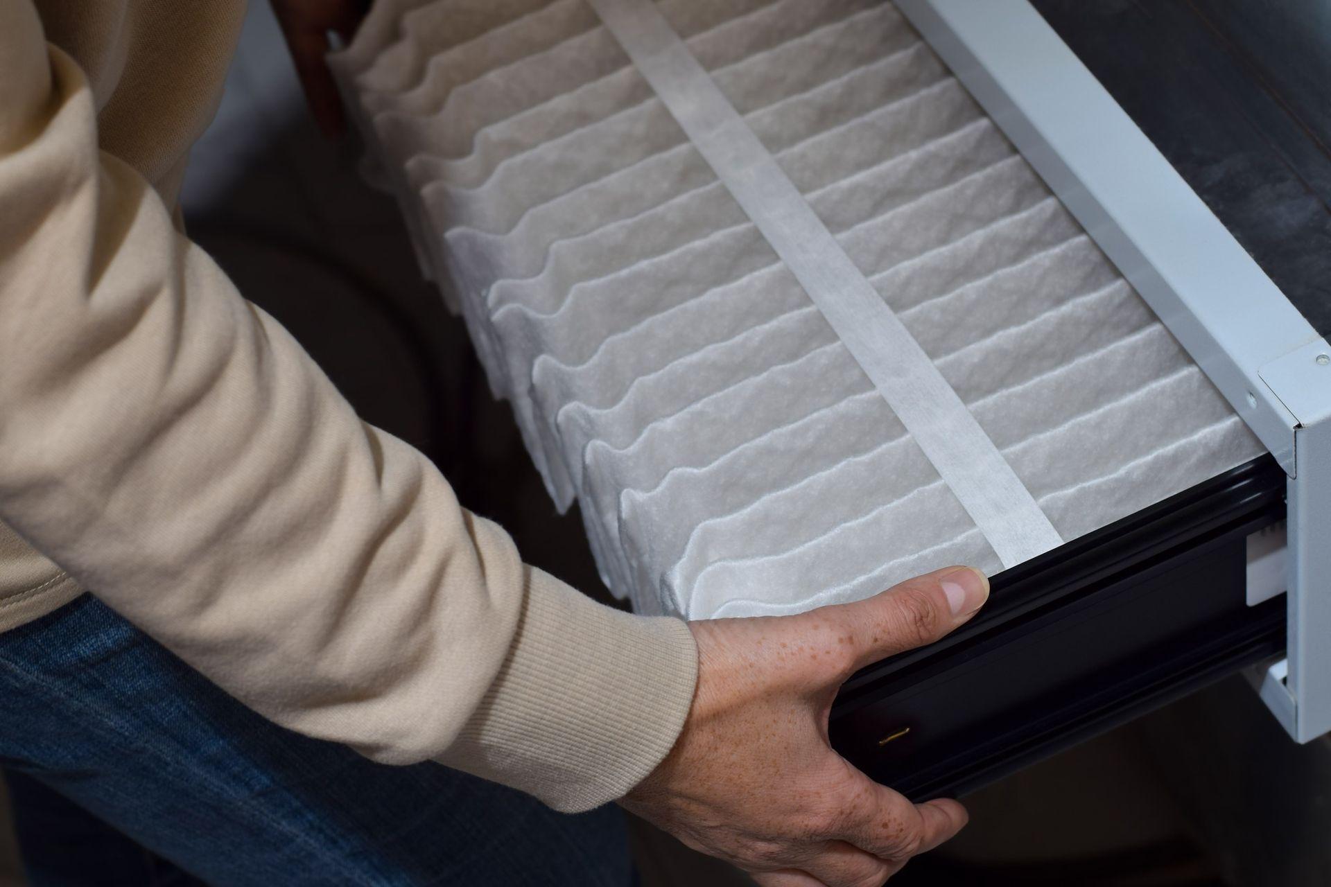 A person in a beige sweater pulls a pleated white air filter out of an HVAC unit.