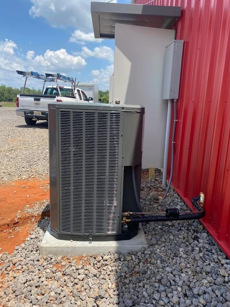 A grey central air conditioning unit sits on a concrete pad outside a red metal building with a service truck nearby.