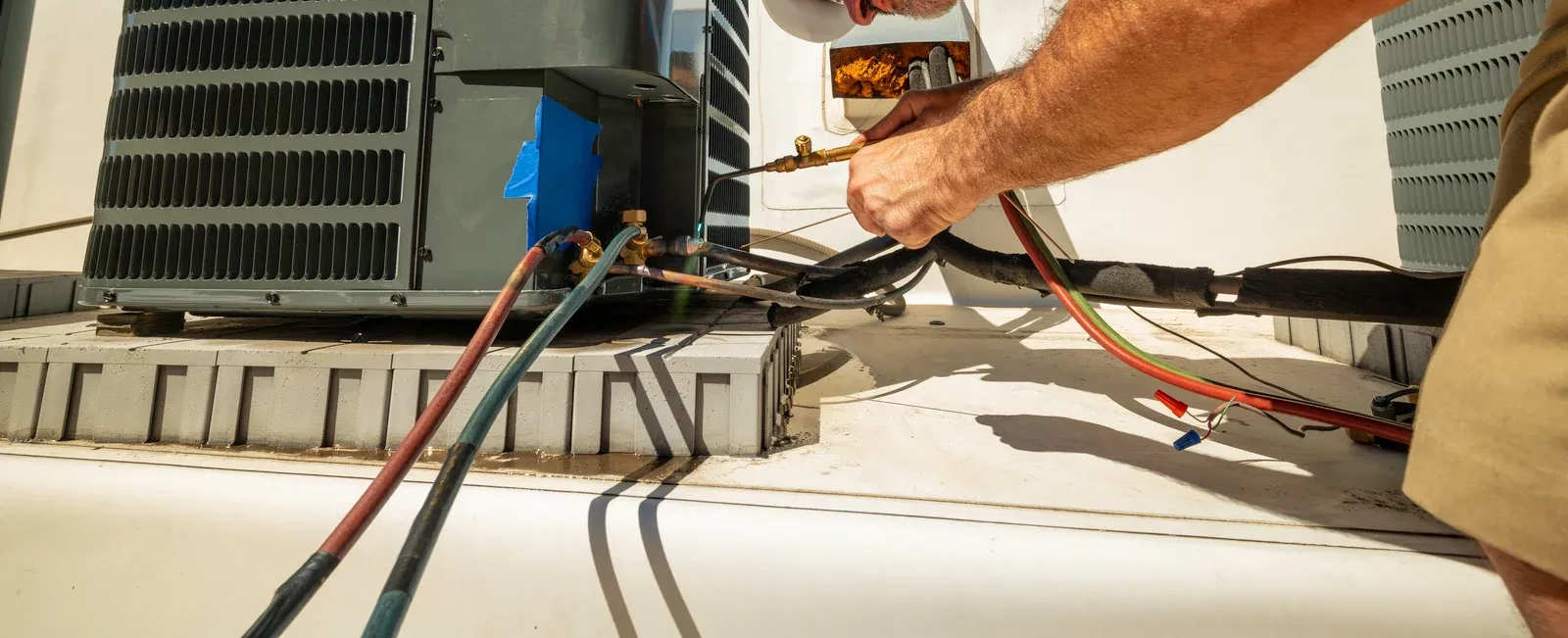 A person in a white shirt and jeans sprays water from a hose onto an outdoor AC unit covered in cleaning foam.