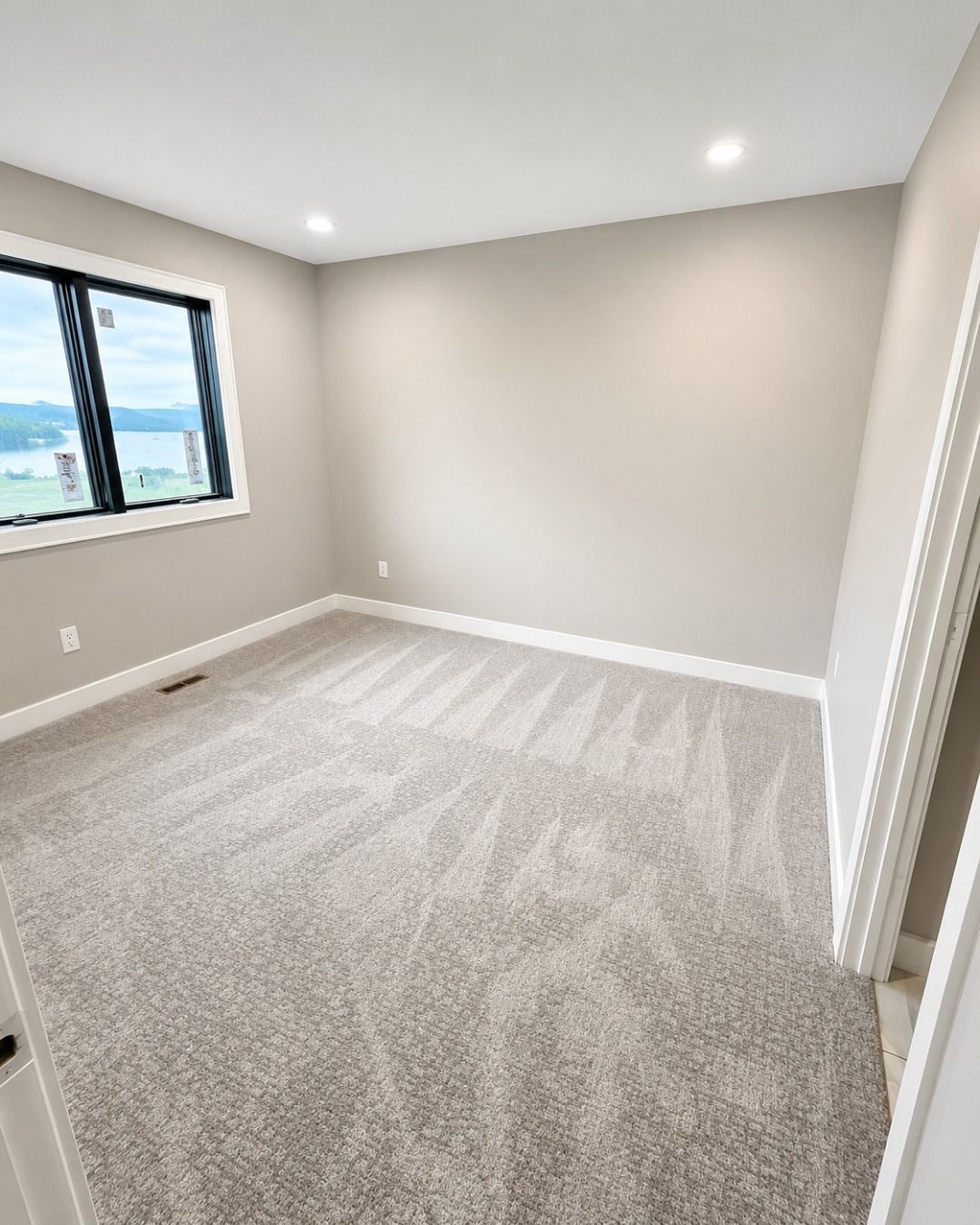 Empty carpeted bedroom with gray walls, recessed lights, and a window on the left.