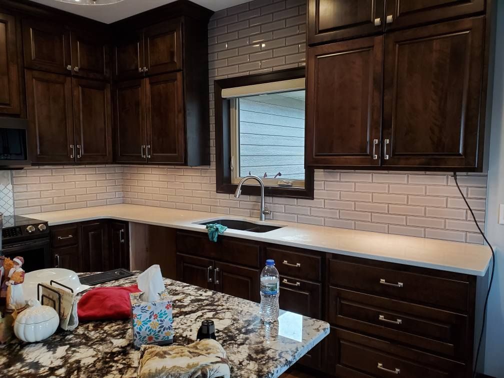 Kitchen with dark wood cabinets, light countertop, brick backsplash, and a sink under a window.