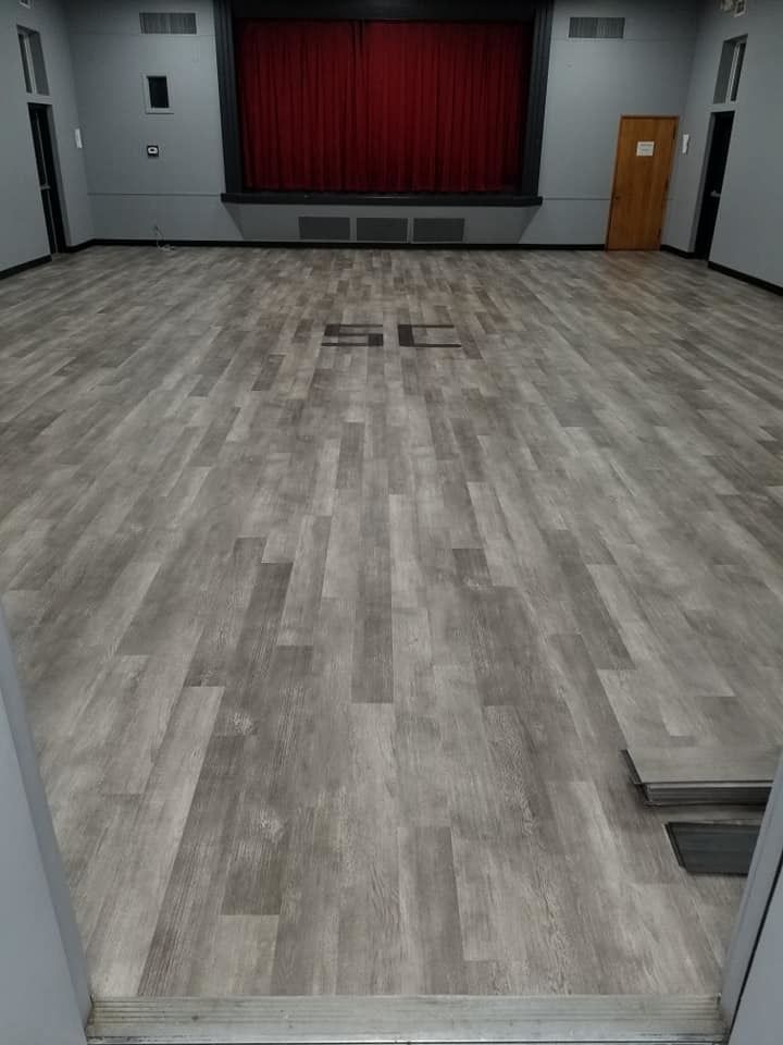 Empty community hall with a small stage, red curtain, and gray wood-patterned floor.