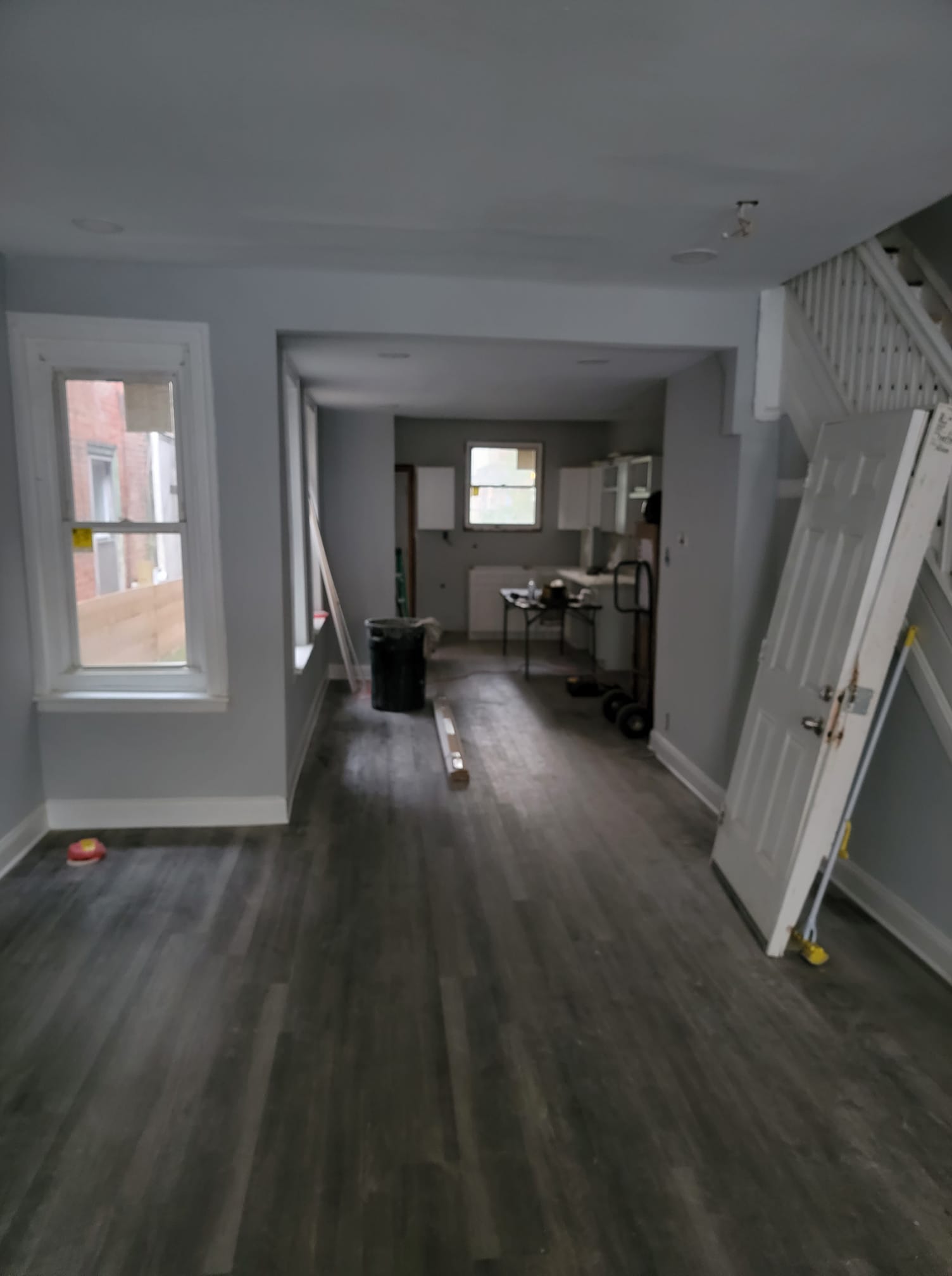 Empty interior room with gray flooring, light blue walls, and unfinished kitchen visible in the background.