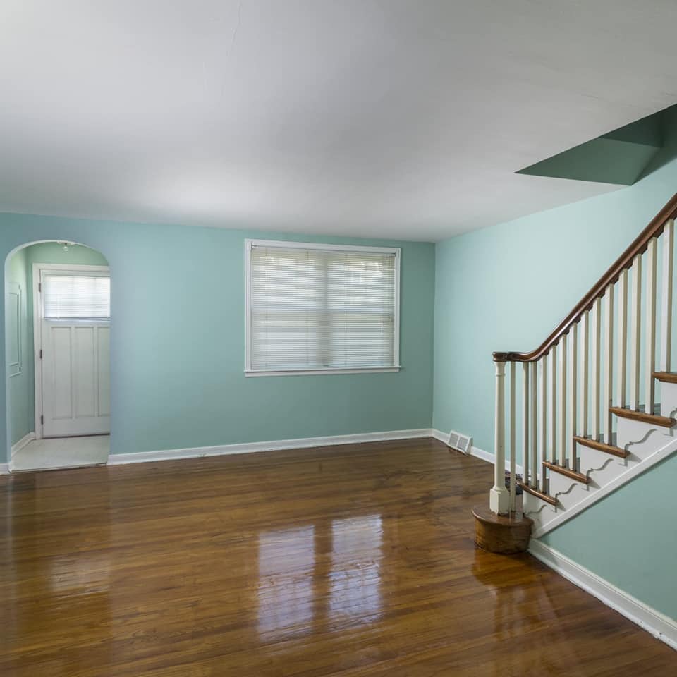 Empty living room with light blue walls, hardwood floors, stairs, and a doorway.