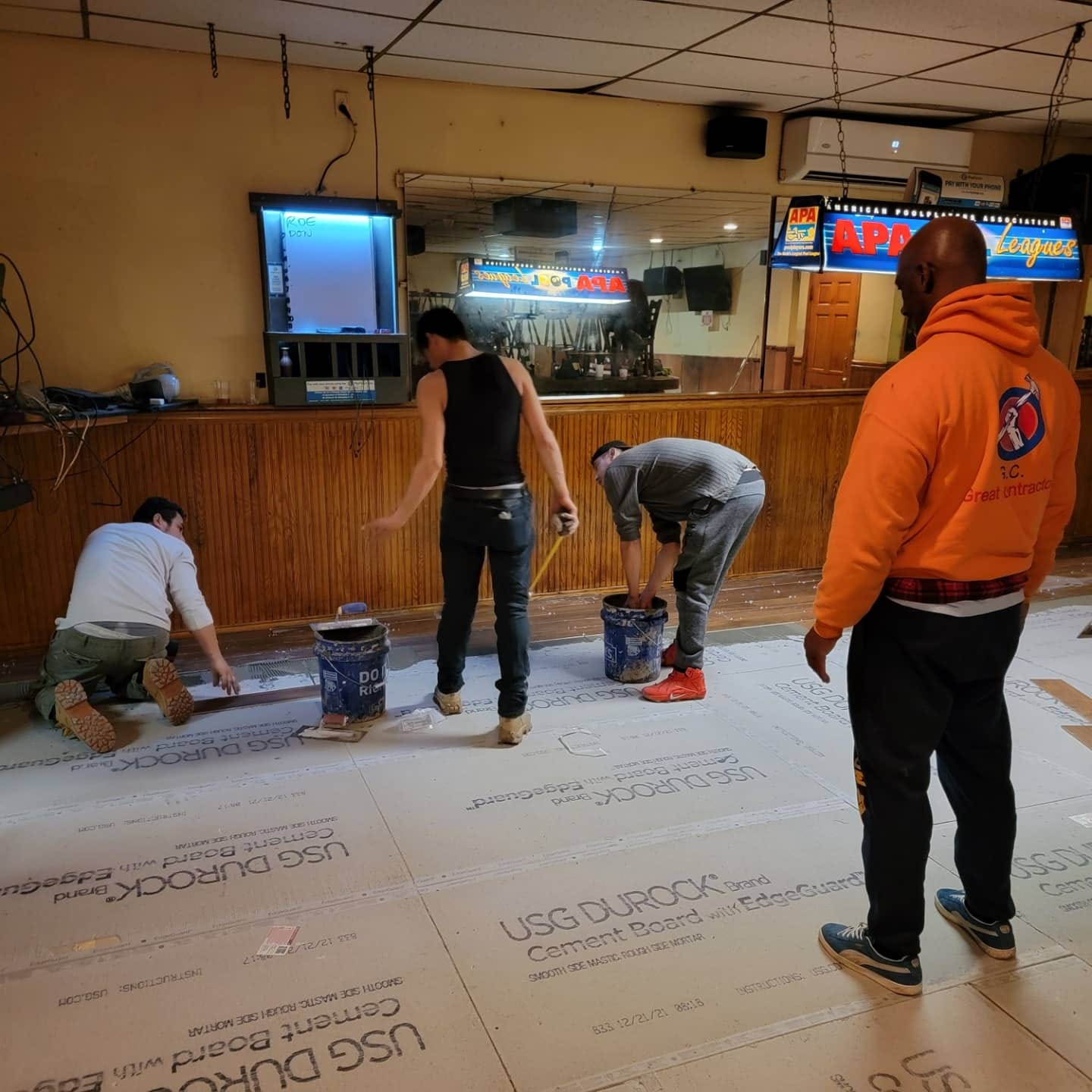 People installing flooring in a bar. Several are standing, kneeling, and working with buckets and cement board.