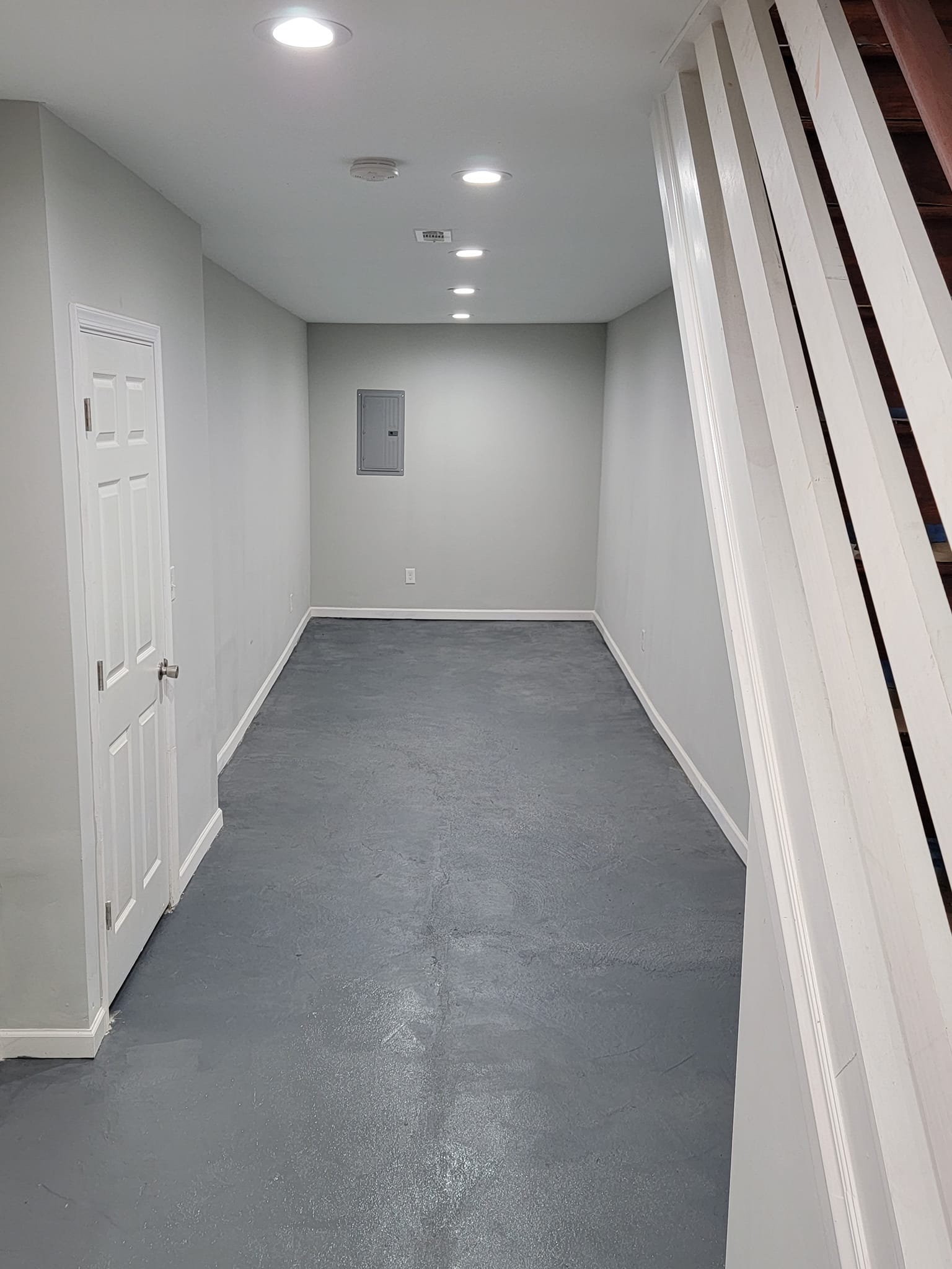 Narrow, gray-walled hallway with a white door, a gray floor, and recessed ceiling lights. A staircase is on the right.