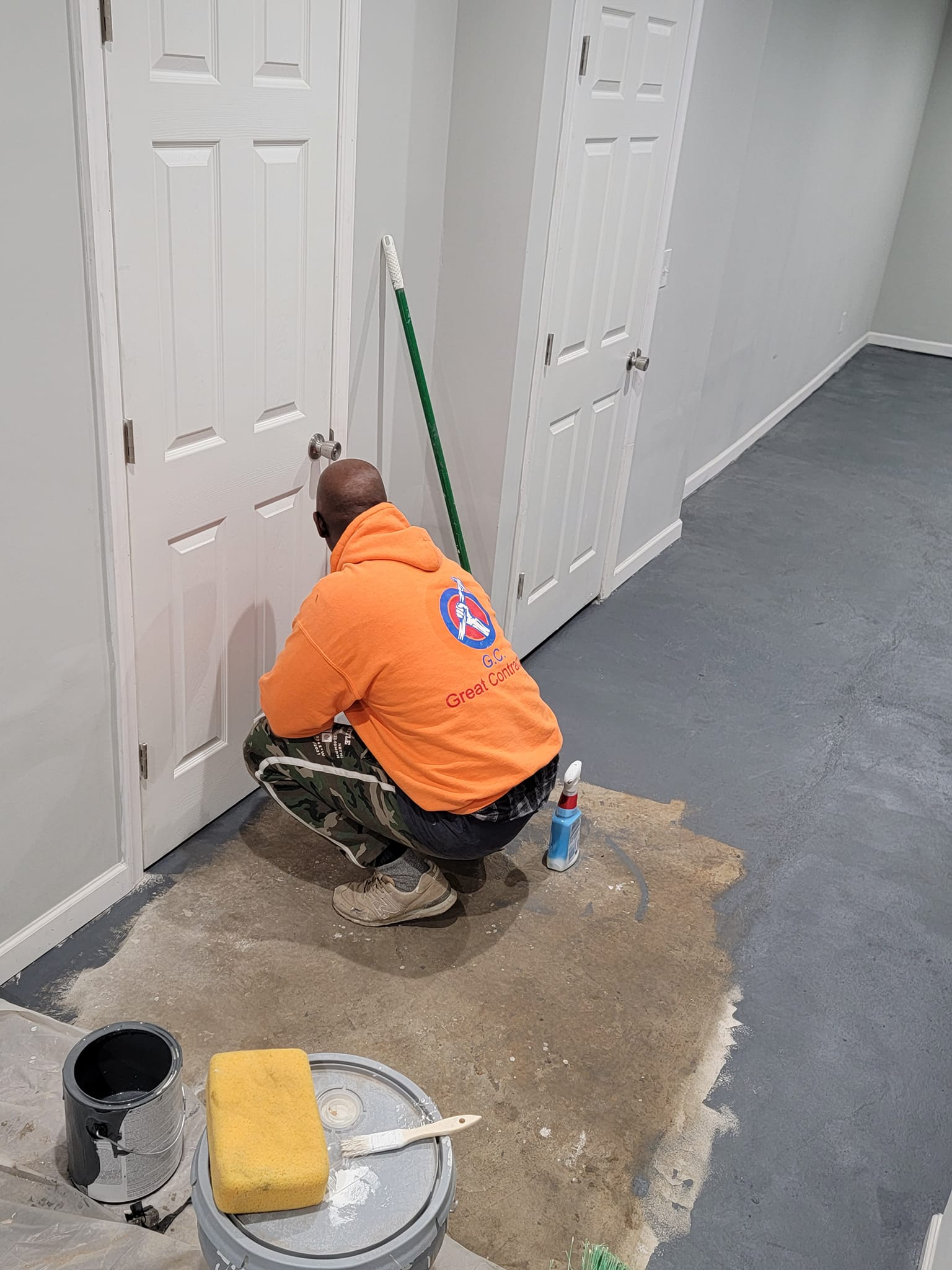 Person painting a white door in a room with gray walls and flooring; paint supplies nearby.
