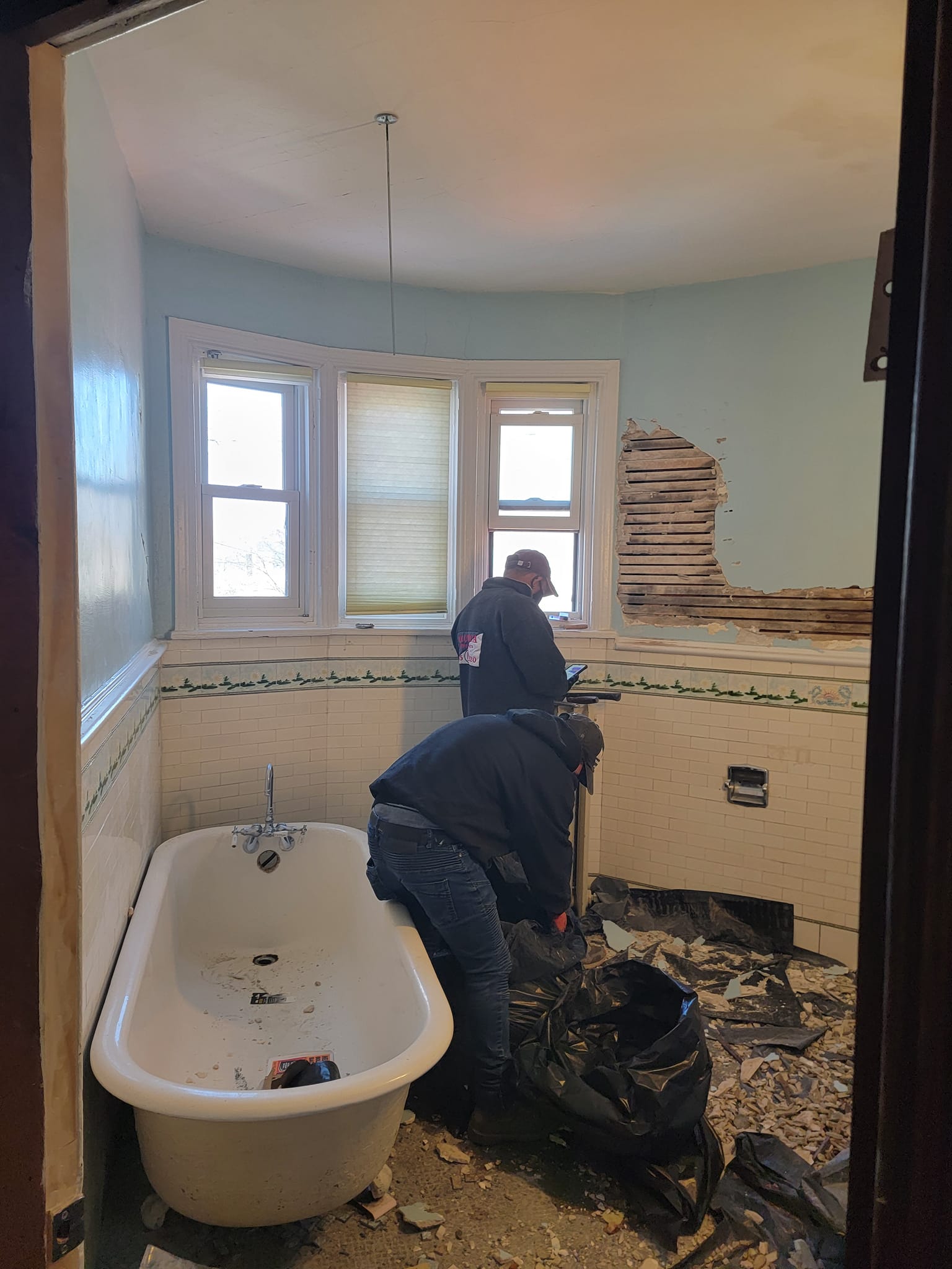 Two people renovating a bathroom with a clawfoot tub, removing drywall.