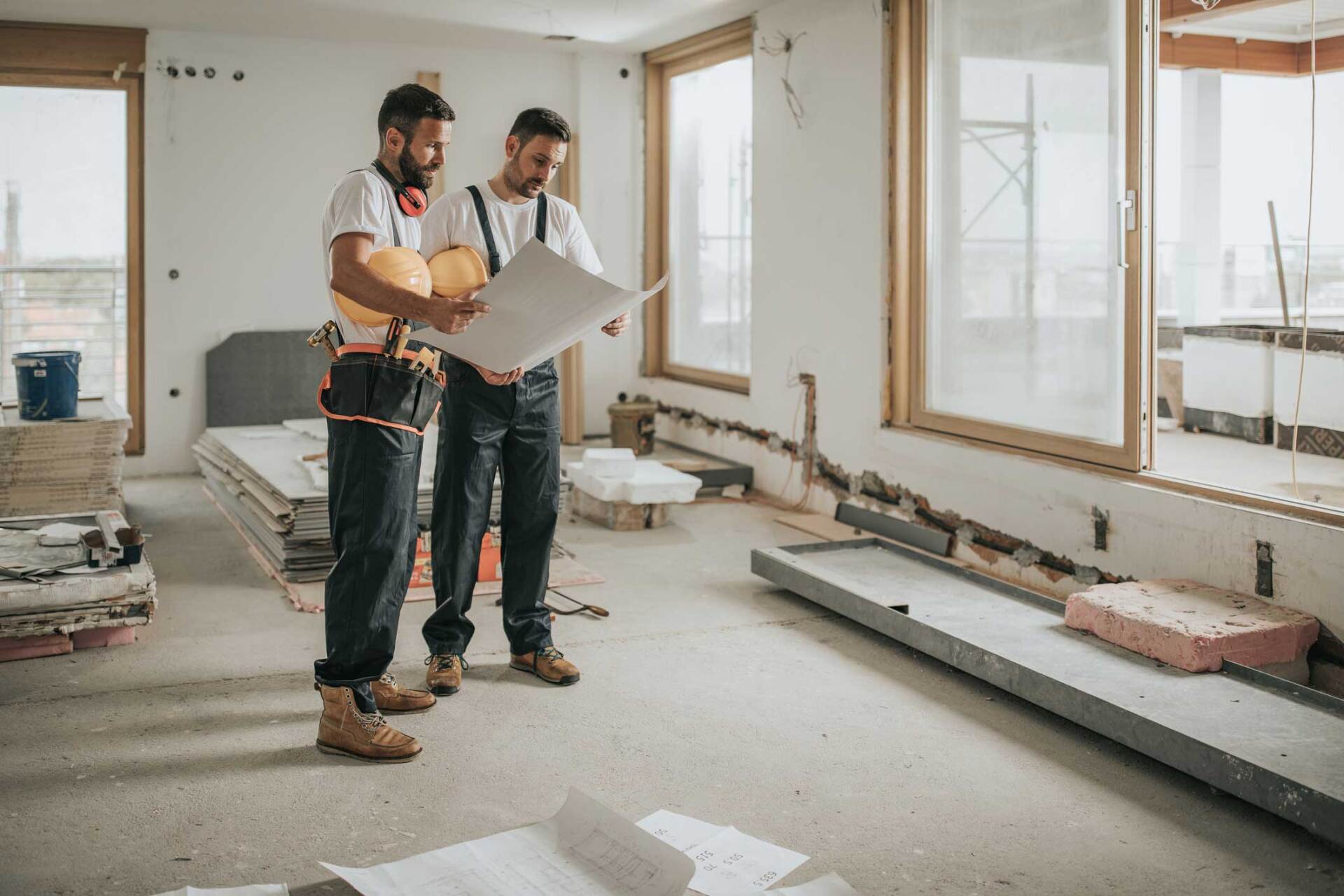 Two construction workers reviewing blueprints in a partially renovated room with windows.