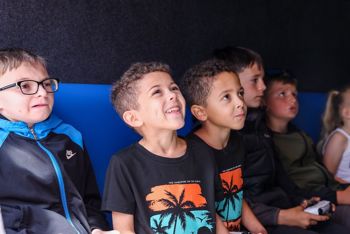 Four children sit in a row, smiling and watching something off-camera indoors.