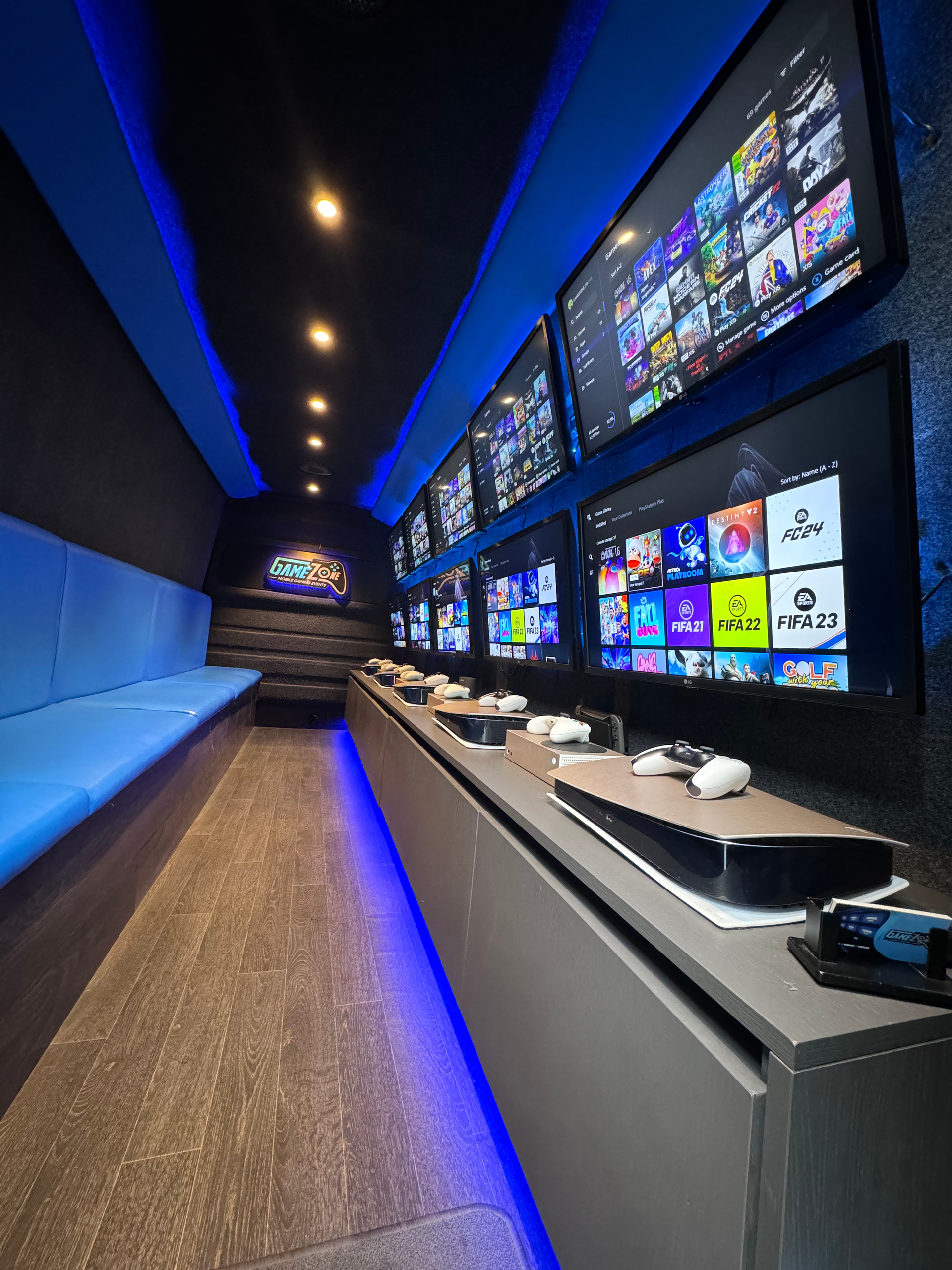 Blue-lit gaming lounge with multiple wall monitors and a long counter of controllers