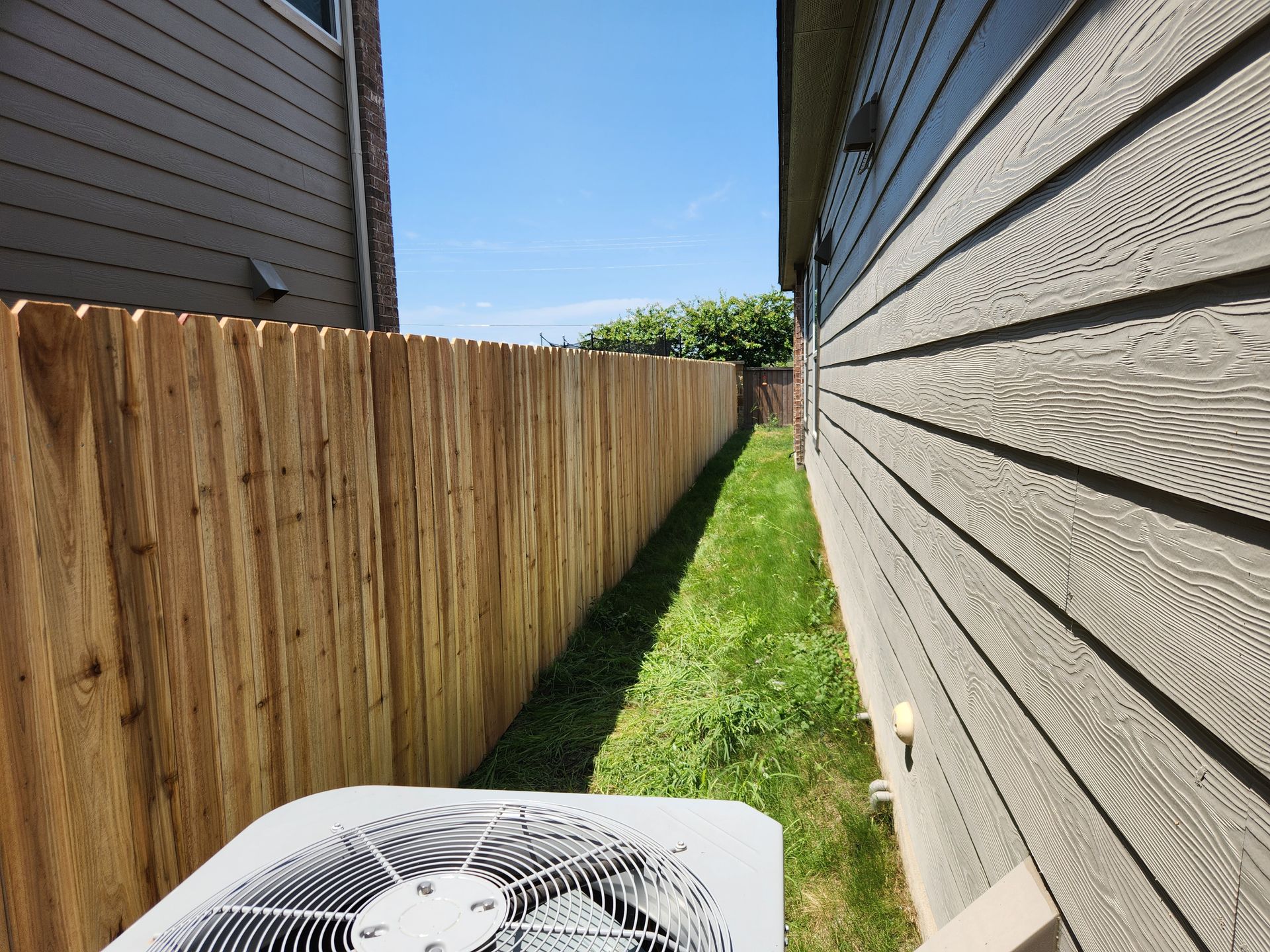 An air conditioner is sitting in the grass next to a wooden fence.
