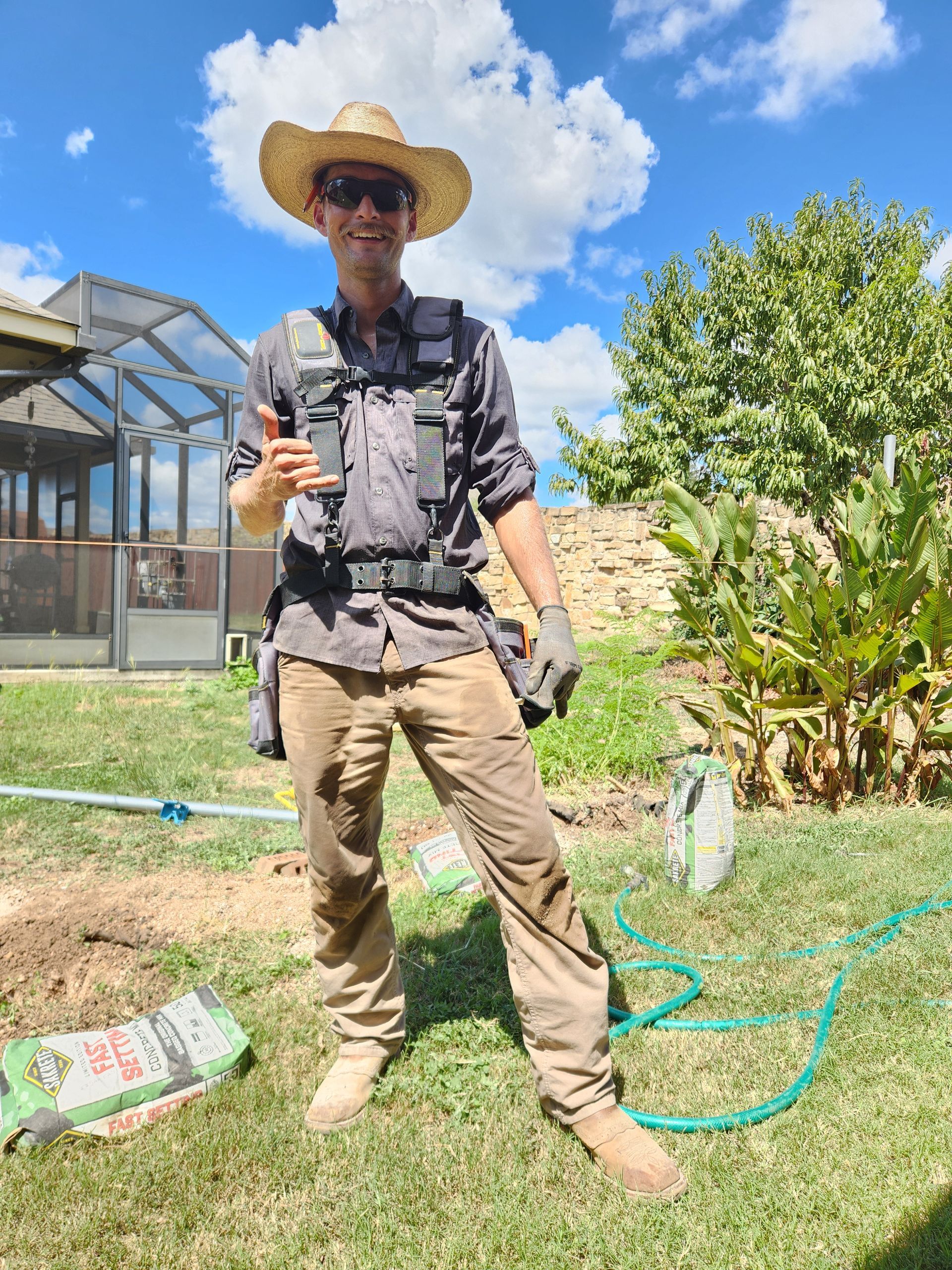 A man wearing a cowboy hat and sunglasses is standing in the grass holding a microphone.