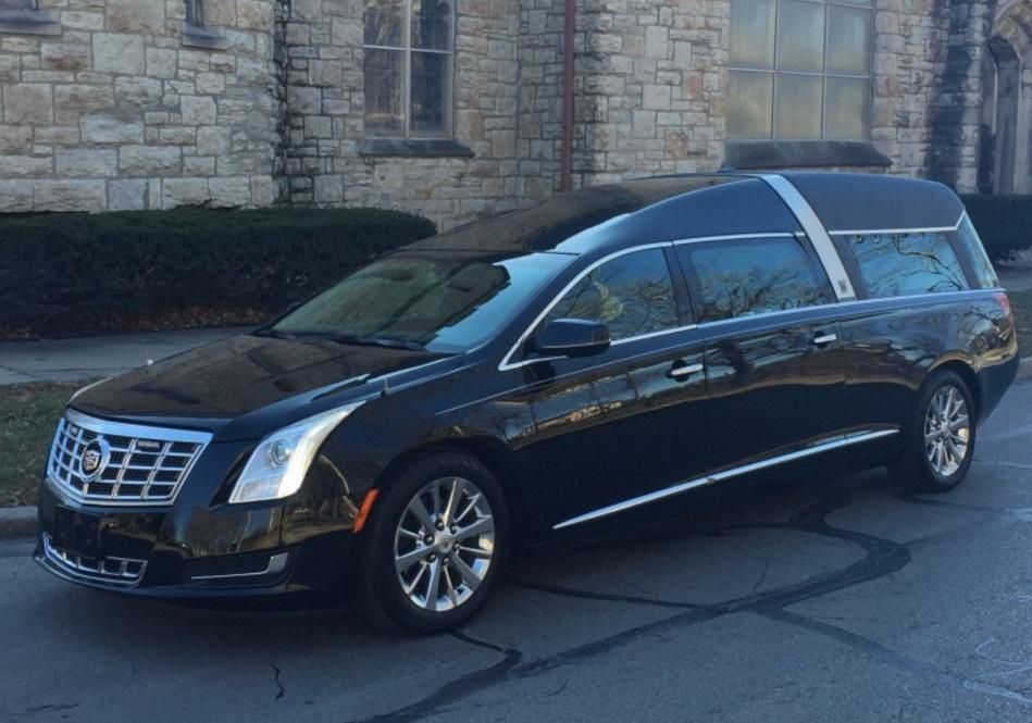 A black funeral car is parked in front of a stone building.