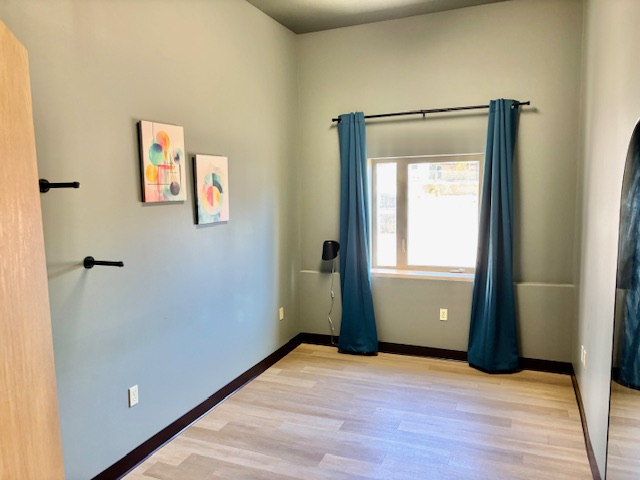 Empty room with blue-gray walls, light wood floor, and teal curtains framing a window. Art and coat hooks on the walls.