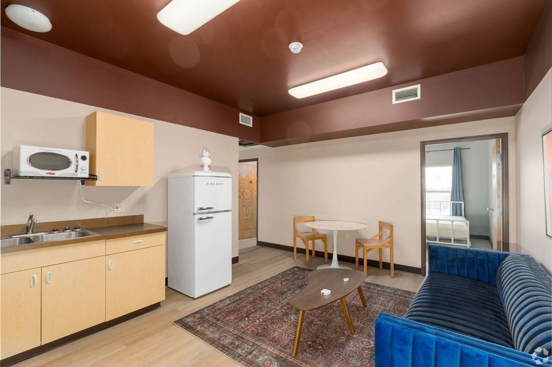 Small apartment with kitchenette, living area, and bedroom. Brown ceiling, blue couch, beige cabinets, and wooden table.