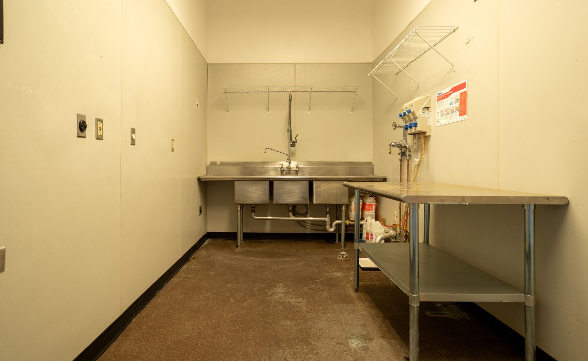 A commercial stainless steel sink and work table in a utility room. Beige walls, concrete floor.