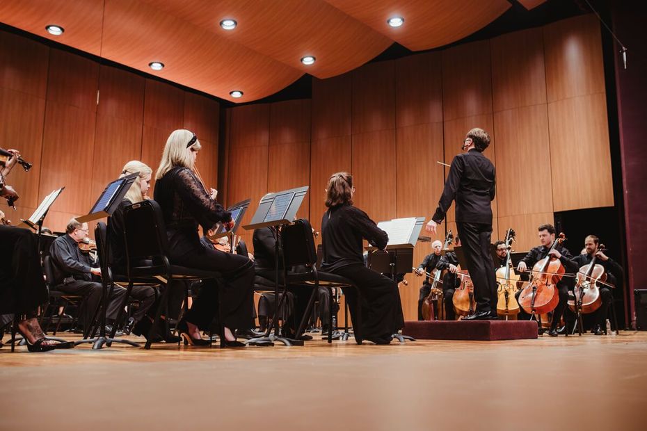 Orchestra performing on a wood-paneled concert hall stage, with conductor leading musicians.