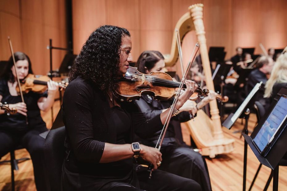Orchestra musicians performing on stage, with a violinist in the foreground and a harp in the background