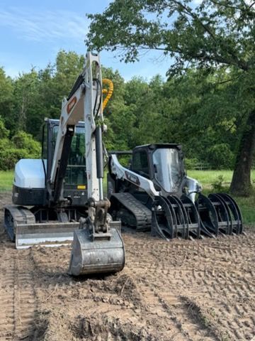 Three construction vehicles parked on dirt in a wooded area, including two mini excavators.