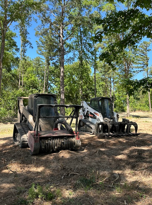 Two skid steer loaders in a clearing with a forest backdrop under a blue sky.
