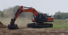 Orange excavator digging in a dusty field with trees in the background