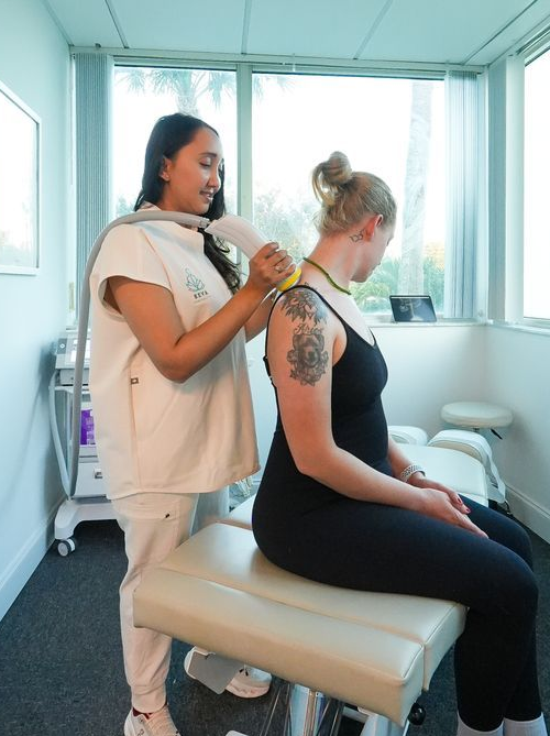Caregiver in white uniform applies therapy device to seated patient’s leg in a clinic