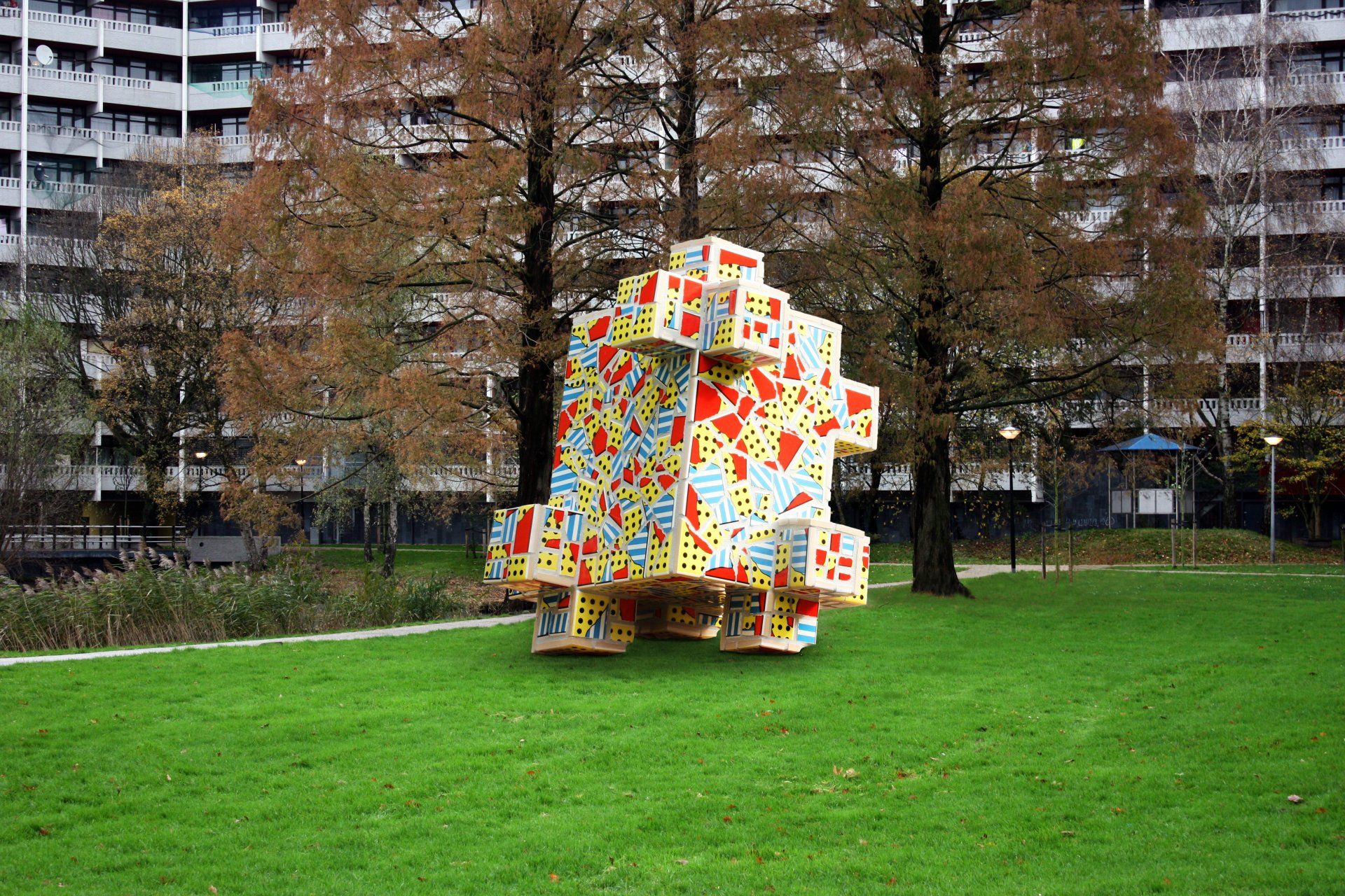 Een kleurrijke geometrische sculptuur op een grasveld met bomen en appartementencomplexen op de achtergrond.