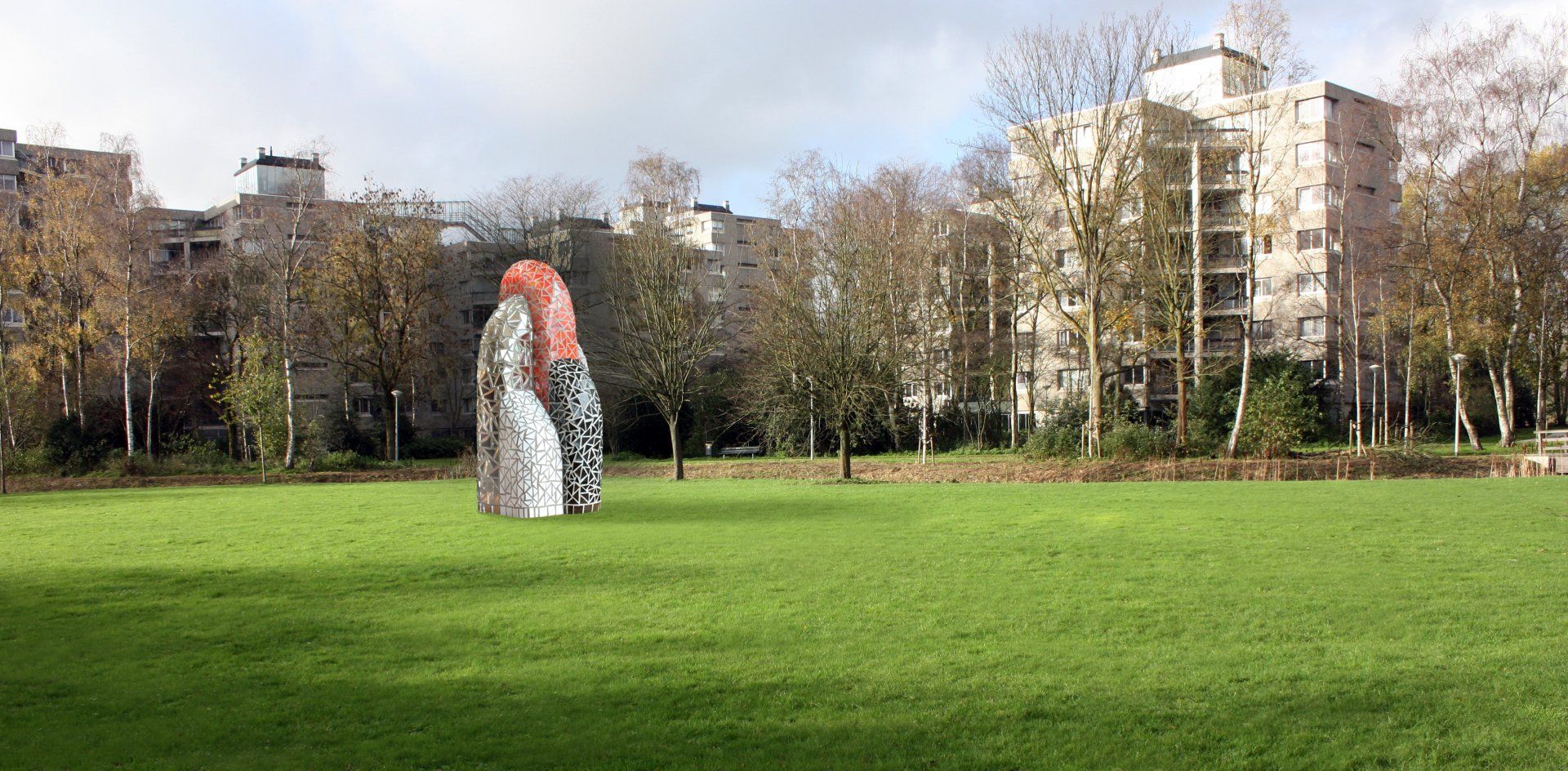 Een grasrijk park met een kleine abstracte sculptuur en herfstbomen voor appartementencomplexen.