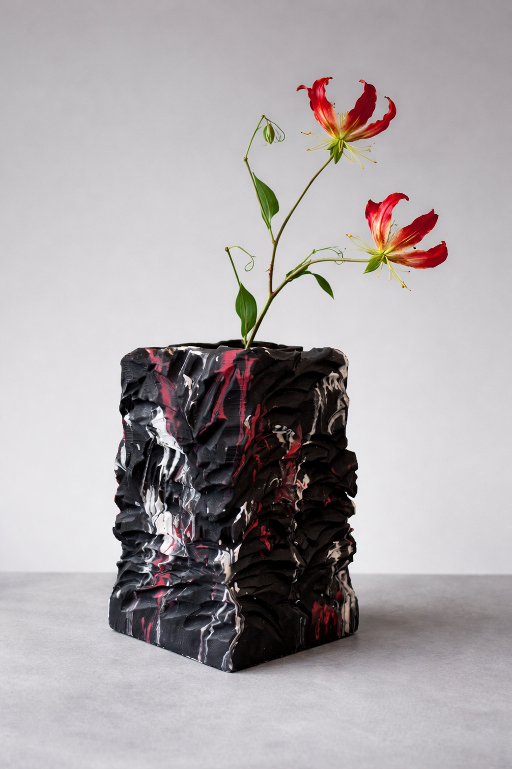 Twee rood-oranje bloemen in een zwarte, gekreukelde vaas op een grijze achtergrond.