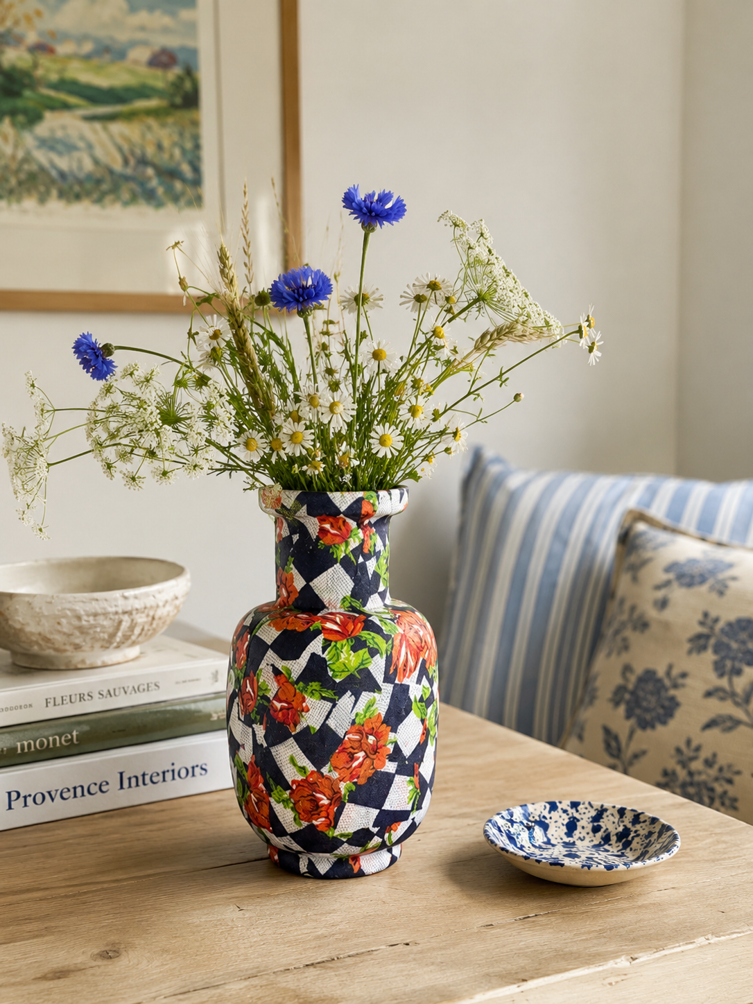 Kleurrijke mozaïekvaas met blauwe bloemen op een houten tafel naast een kom met patroon en een gestreept kussen.