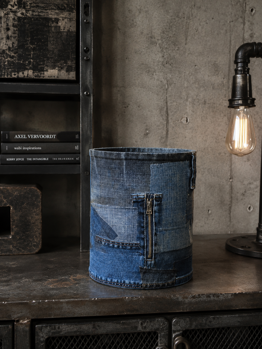 Een blauwe denim emmer staat op een donkere plank naast boeken en een brandende lamp in industriële stijl.