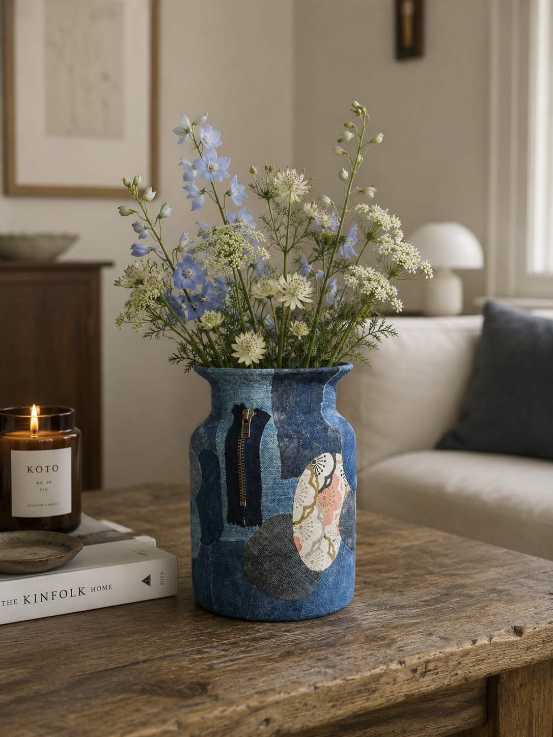 Blauwe keramische vaas met wilde bloemen op een rustieke houten tafel naast een brandende kaars en boeken.