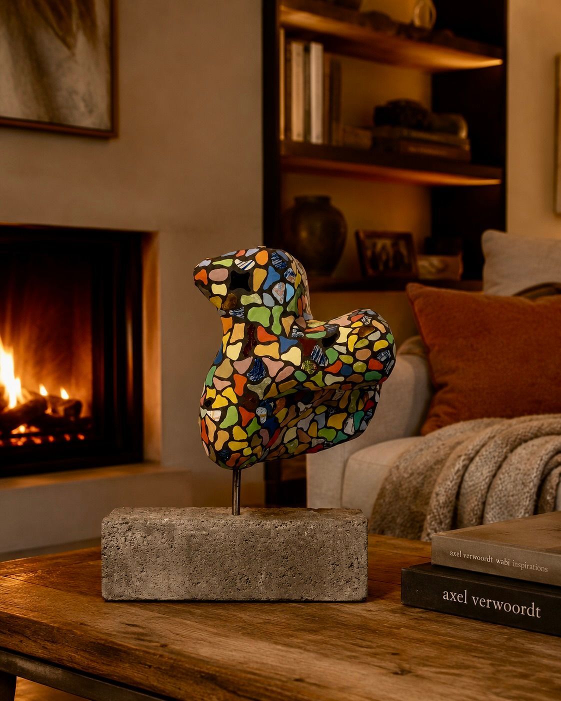 Mozaïek vogelsculptuur op een stenen sokkel naast een brandende open haard in een gezellige woonkamer.