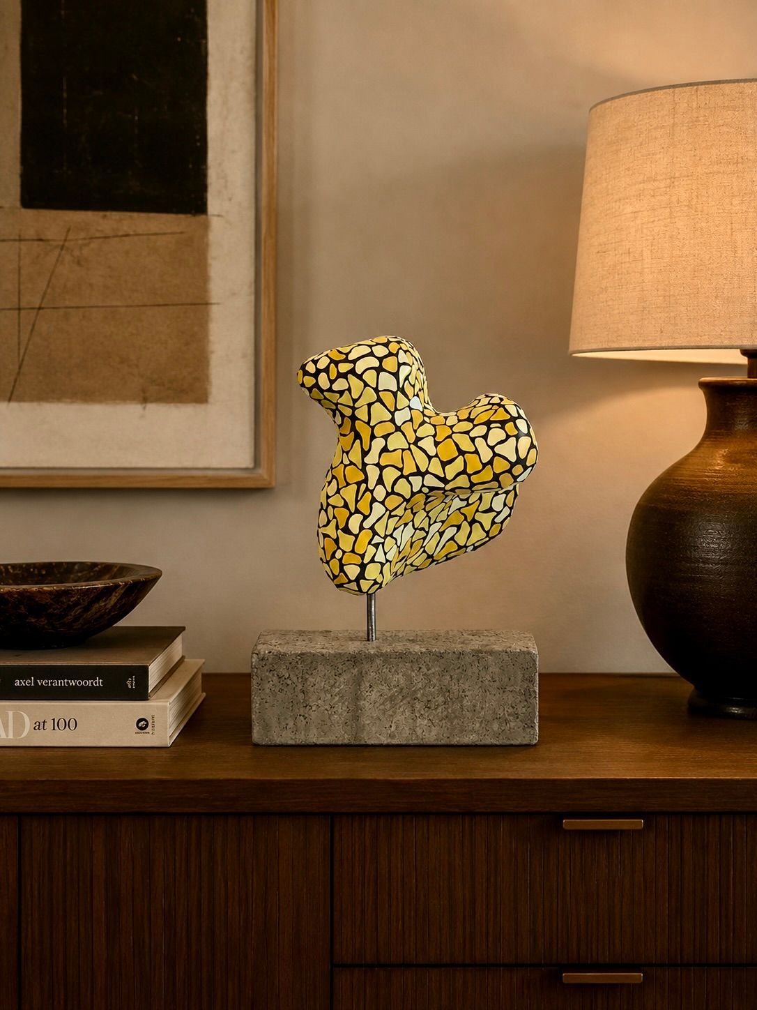 Abstracte geel-zwarte figuur op een houten console naast een lamp en opgestapelde boeken.