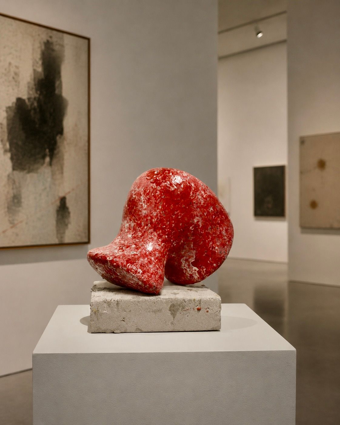 Een rode abstracte sculptuur op een witte sokkel in een galerie, met ingelijste kunstwerken aan de muren.