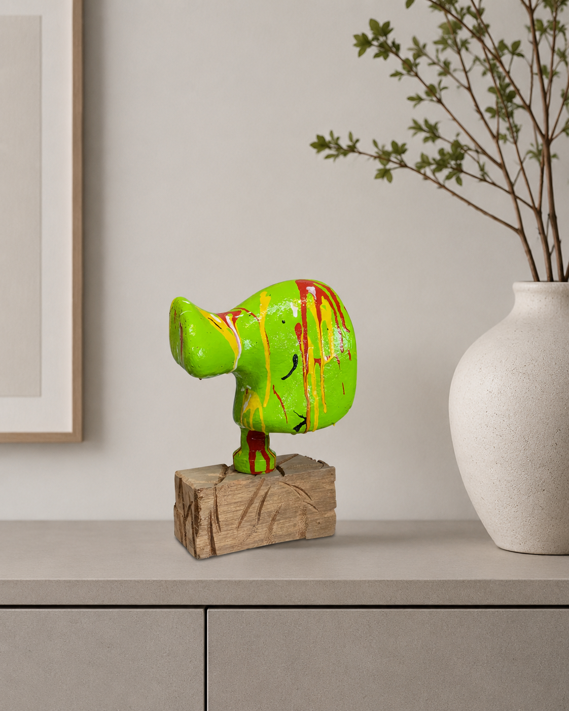 Neongroene abstracte sculptuur met rode druppels op een houten sokkel naast een vaas op een tafel.