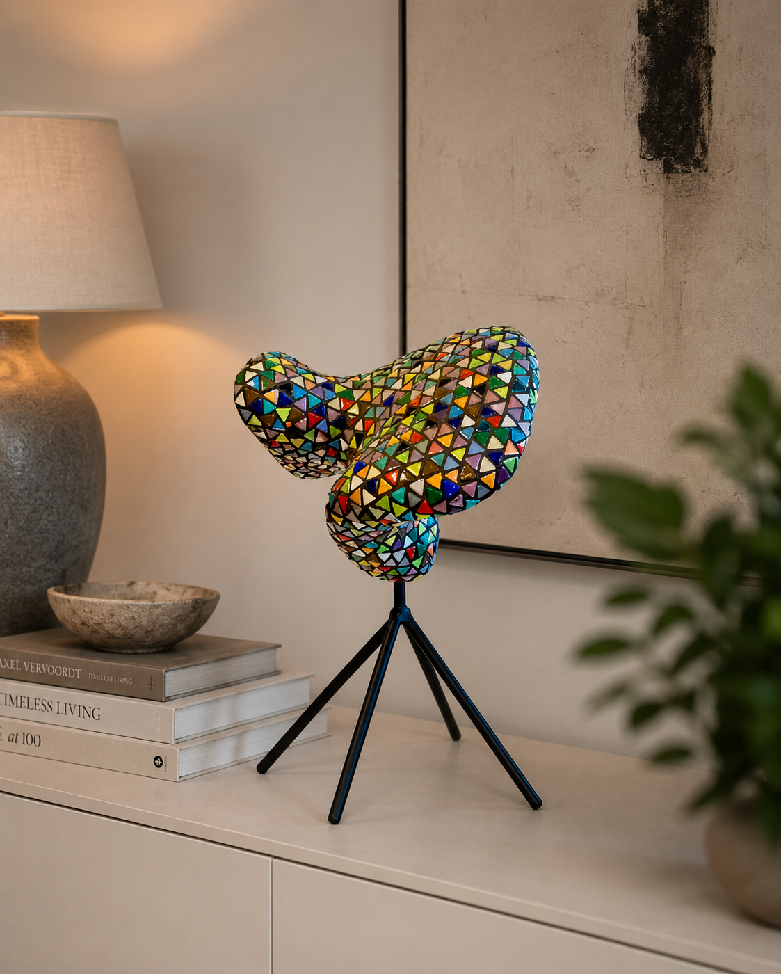 Een kleurrijke mozaïeksculptuur in de vorm van een vlinder, op een zwarte sokkel naast een lamp en boeken op een plank.