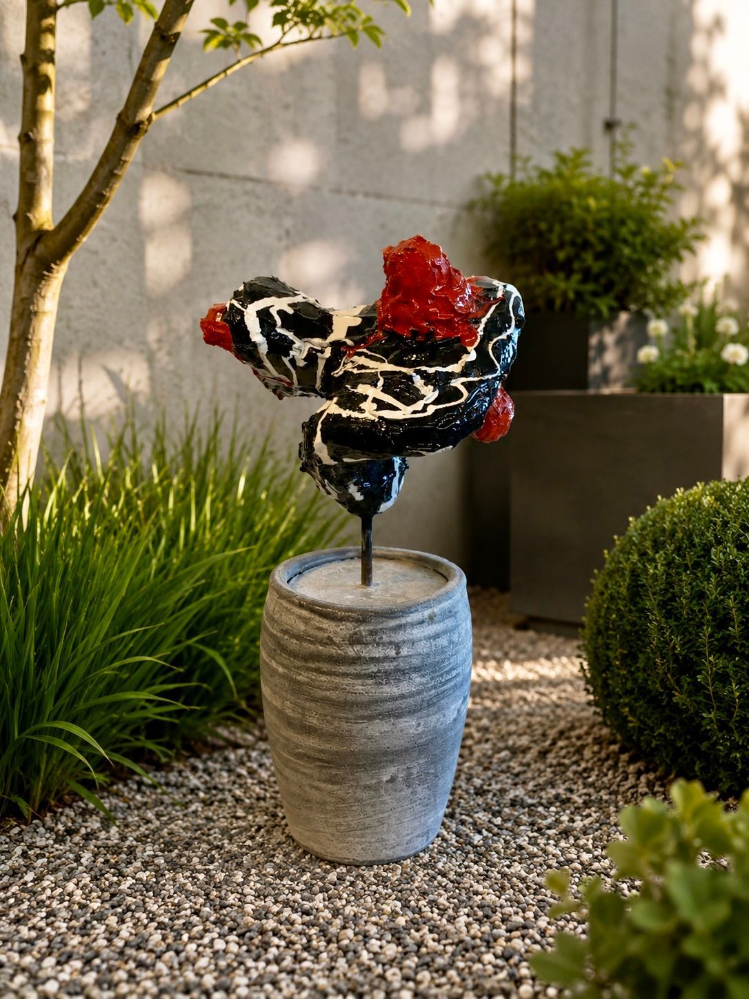 Abstracte sculptuur in zwart, wit en rood in een grijze plantenbak op een grindpad in de tuin.