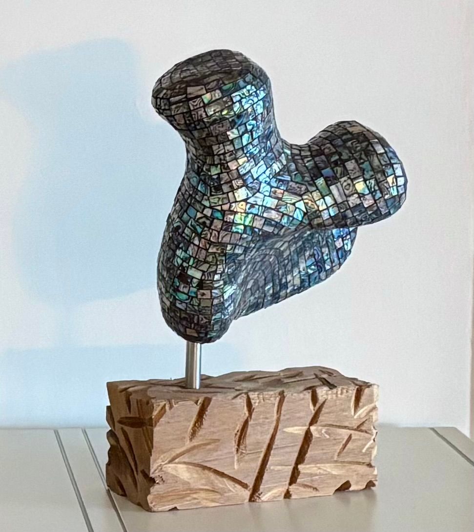 Abstracte mozaïeksculptuur op een houten sokkel tegen een effen lichte achtergrond.