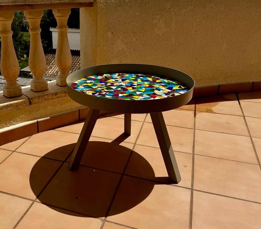 Ronde kruk met een kleurrijk mozaïekblad op een zonnig betegeld terras.