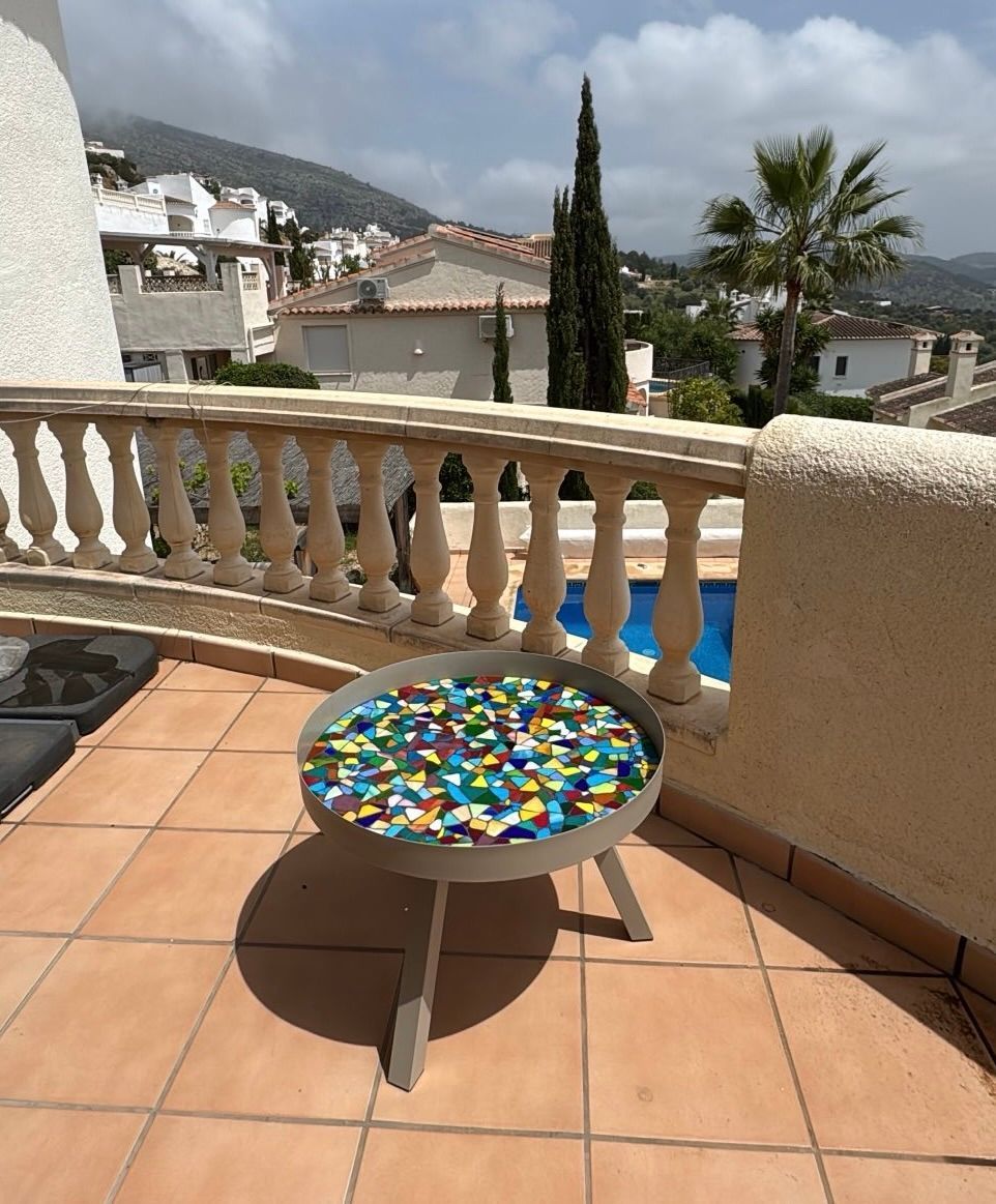 Terras met een kleurrijk mozaïek tafelblad, terracotta tegels en een witte balustrade met uitzicht op heuvels en palmbomen.